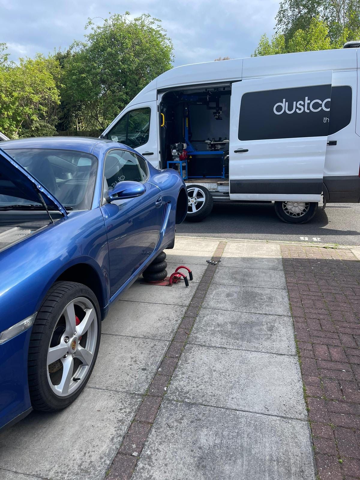 Blue Porsche jacked up on a driveway with our white van and its workshop open behind